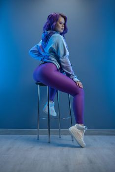 girl in stylish purple sportswear and with purple hair poses sexually on a bar stool