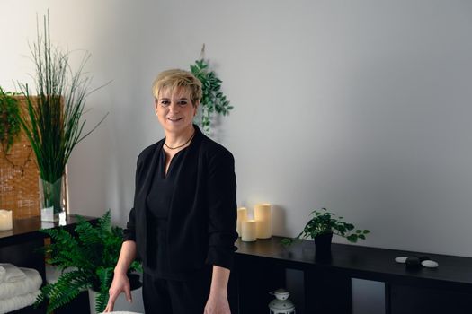 Portrait of a mature, smiling female beautician, dressed in black company uniform, posing standing in her small spa business, to start the day. Relaxing atmosphere and subdued lighting, decoration of plants and candles, background massage table and towels. Horizontal