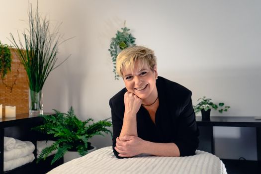 Portrait of a mature, smiling female beautician, dressed in black company uniform, posing standing in her small spa business, to start the day. Relaxing atmosphere and subdued lighting, decoration of plants and candles, background massage table and towels. Horizontal