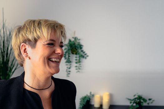 Portrait of a mature, smiling female beautician, dressed in black company uniform, posing standing in her small spa business, to start the day. Beautician leaning smiling on a massage table in her spa business. Relaxing atmosphere and subdued lighting, decoration of plants and candles, background massage table and towels. Horizontal. copy space on the right