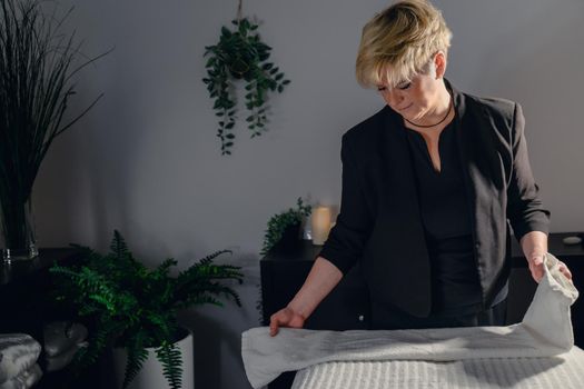 Portrait of a blonde-haired woman, beautician, mature, concentrated and professional, dressed in the black uniform of the company, preparing the massage table to start the day. Relaxing atmosphere and subdued lighting, decoration of plants and candles, background massage table and towels. Horizontal.