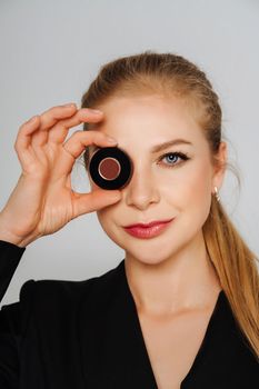 A beautiful middle-aged woman covered one eye with a round box and smiled. Blond hair and a black jacket on a light background