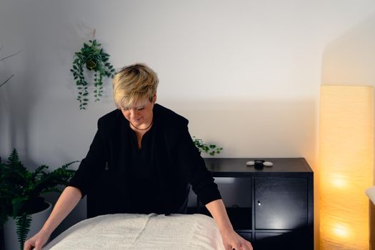 Portrait of a blonde-haired woman, beautician, mature, concentrated and professional, dressed in the black uniform of the company, preparing the massage table to start the day. Relaxing atmosphere and subdued lighting, decoration of plants and candles, background massage table and towels. Horizontal.