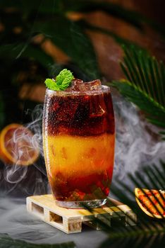 Bumble coffee with ice on a dark tropical background. Espresso, orange juice and syrup in layers in transparent glass with ice. Summer trendy refreshing coffee drink. Coffee house menu, recipe. Copy space