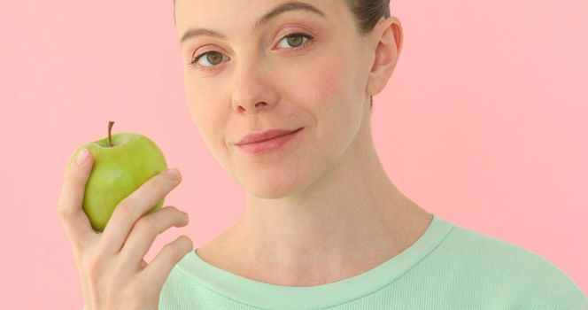 Woman enjoying fresh apple at pink background. Girl looking at camera