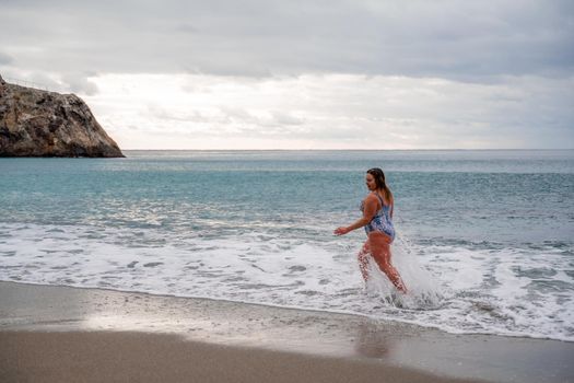 A plump woman in a bathing suit enters the water during the surf. Alone on the beach, Gray sky in the clouds, swimming in winter