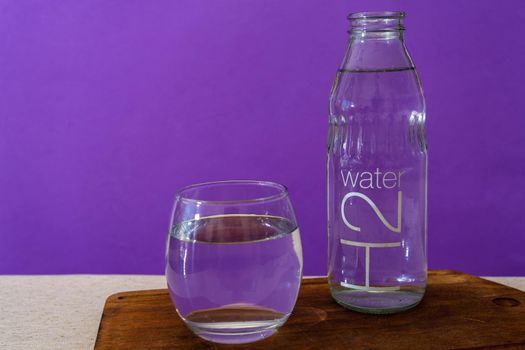 Glass beaker and bottle filled with purified water. Violet background.