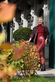 fashionable blonde girl with a red lipstick posing outdoors . Dressed in a red leather leggings, turtleneck and checkered jacket. low lace up square toe block heel ankle black leather boots.