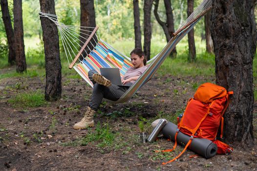 Caucasian woman working on laptop while sitting in a hammock in the forest. Girl uses a wireless computer on a hike