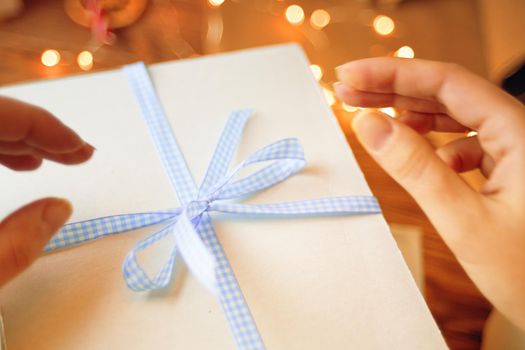 Close-up of women's hands and blue checkered ribbon on white gift box. Christmas or Birthday surprise. Garland lights on background.