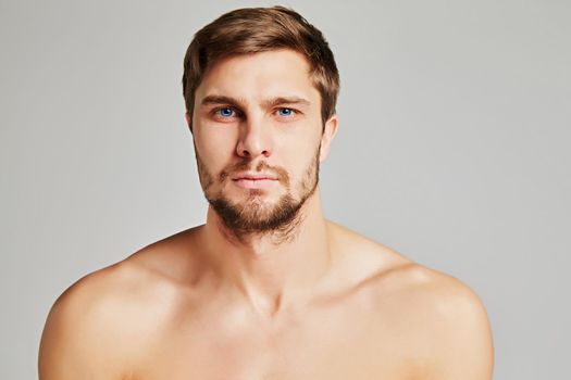 Portrait of a serious young man with bare shoulders on a gray background, powerful swimmers shoulders, beard, charismatic, adult, brutal, athletic. High quality photo