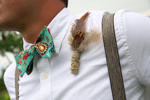 Close up of a Feather and Burlap Boutonniere and Turquoise Paisley Bowtie Worn by Man in White Dress Shirt and Suspenders. High quality photo