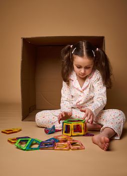 European little girl with two ponytails in pajamas playing with colorful magnetic constructor toy on beige background with copy ad space