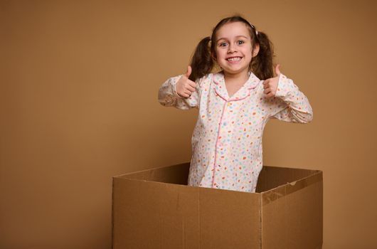 Charming cheerful child, pretty Caucasian baby girl with two ponytails showing thumbs up, smiles looking at camera being inside a cardboard box against a beige background with copy advertisement space