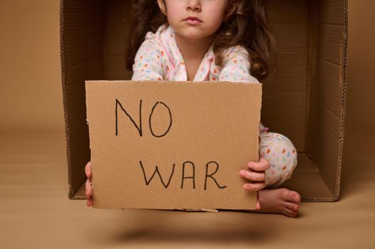 No War written in a cardboard poster in the hands of European child little girl refugee or migrant during political and military conflicts in the country, isolated over beige background, copy ad space