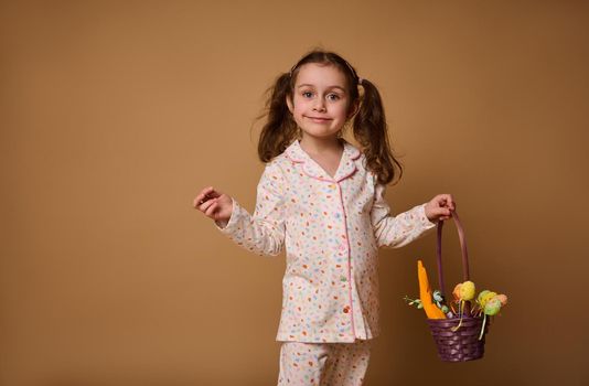 Portrait of a beautiful Caucasian child girl in pajamas with bright dots stands with a purple basket and colored Easter eggs, isolated on a beige background with space for advertising text