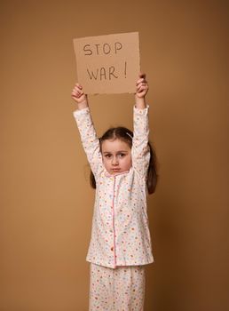 Studio shot of an adorable upset 4 years old kid, sad little European girl in pajamas holding Stop War social message cardboard poster standing on beige background with ad copy space