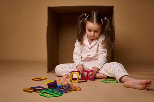 Charming child, European little girl concentrated on building with colorful magnetic constructor, developing her fine motor skills, isolated over beige background with copy space