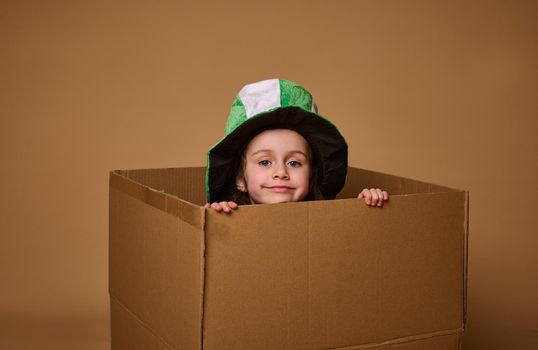 Adorable 4 year old child girl with a Leprechaun hat peeks out from under a cardboard box and smiles cutely at the camera. Happy St. Patrick's Day. Studio shot on beige background with copy space