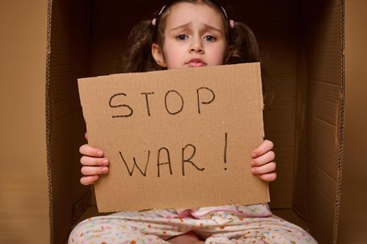 Close-up little caucasian girl in home clothes sitting inside a cardboard box with a STOP WAR poster. The concept of refugees and migrants who lost their homes during political and military conflicts