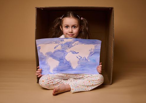 Adorable Caucasian child 4 years old girl sitting inside a cardboard box with a poster- painted image with World map, isolated over beige background.Environment conservation and save planet concept