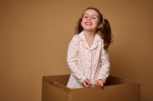 Cheerful little European girl with two wavy ponytails, wearing white pajamas with colorful dots, smiling with a beautiful toothy smile, standing inside a cardboard box, isolated over beige background
