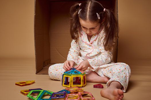 Concentrated little preschool girl, Caucasian child in pajamas sitting ahead a cardboard box and playing with magnetic constructor. Kids entertainment and fine motor kills development