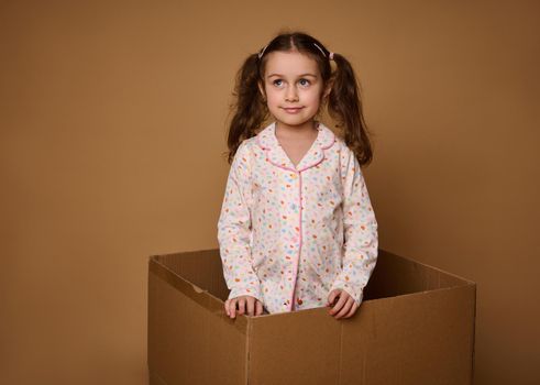 Adorable European 4 years old baby girl with two wavy ponytails wearing a bright pajamas looking aside while standing inside a cardboard box, isolated over beige background with copy space for ads