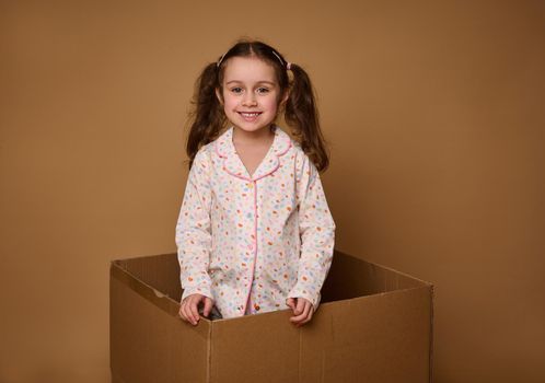 Adorable child, European 4 years old baby girl with two wavy ponytails in a bright pajamas looking at camera standing inside a cardboard box, isolated over beige background with copy space for ads