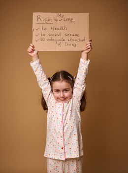 Adorable European preschool girl holding cardboard poster promoting children's rights to an adequate standard of living, social secure and health care, isolated on a beige background with copy space