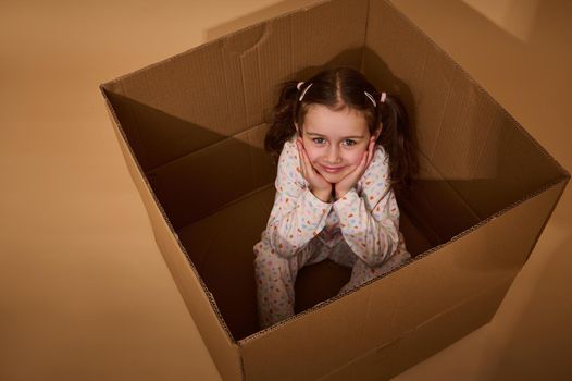 Overhead view of a charming little Caucasian girl in pajamas with colorful dots sitting inside a cardboard box and looking at camera, isolated over beige background with copy ad space