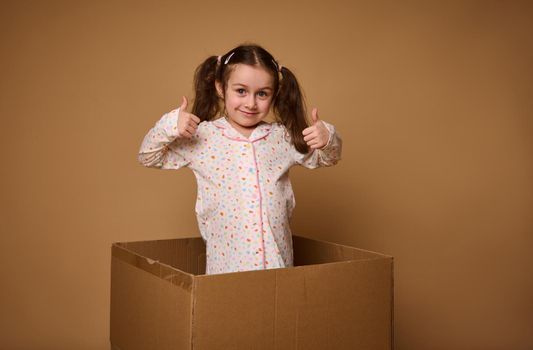 Charming cheerful kid, pretty Caucasian baby girl with two ponytails showing thumbs up, being inside a cardboard box against a beige background with copy ad space