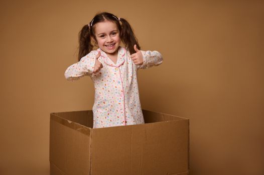 Adorable Caucasian little girl, cheerful kid with two ponytails showing thumbs up, smiles looking at camera being inside a cardboard box against a beige background with copy advertisement space