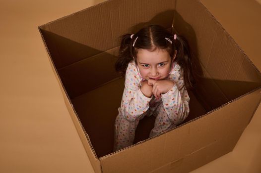 Studio shot of a sly beautiful little girl looking mysteriously at the camera while inside a cardboard box. Emotional studio portrait on a beige background with space for advertising