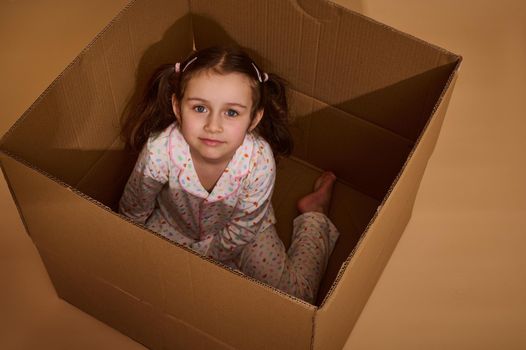 Top view of a charming little Caucasian girl in pajamas with colorful dots sitting inside a cardboard box and looking at camera, isolated over beige background with copy ad space