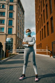 Full length shot of sporty woman in sportswear, headphones and medical mask standing on a street