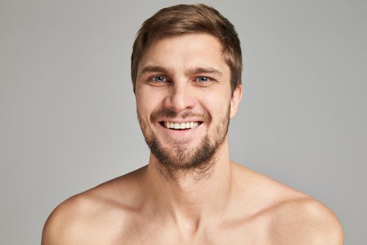 Portrait of a smiling young man with bare swimmers shoulders on a gray background, powerful, beard, charismatic, adult, brutal, athletic, edited photo, bright smile, white teeth smile, look in camera. High quality photo