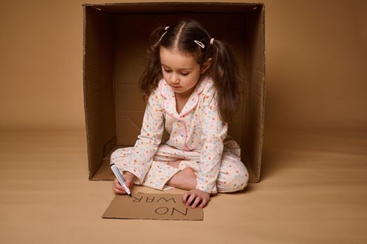 Cute child, adorable Caucasian kid girl in home clothes hiding from war inside cardboard box, writing social message- Stop War. Concepts of refugees migrants losing their home during military conflict