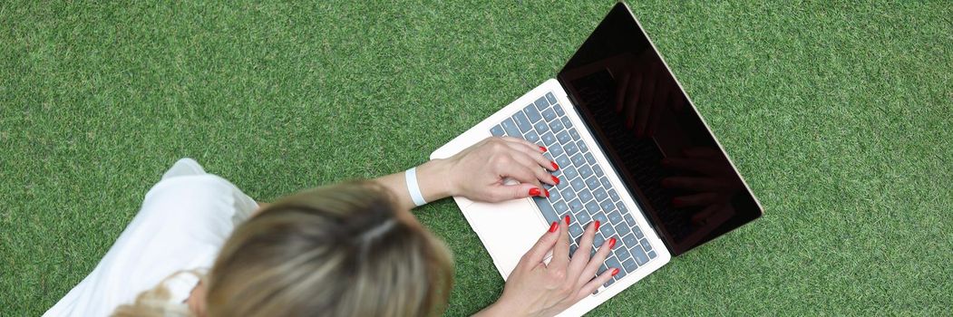Young woman lying on green grass and typing on laptop keyboard top view. Communication on social networks concept