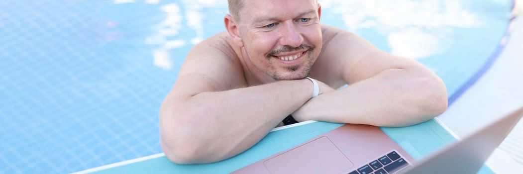 Man lying on side of swimming pool and looking at laptop screen. Remote work at resort concept