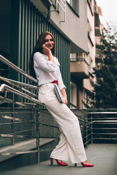 Beautiful young caucasian businesswoman in a white clothes with a laptop and notebook outdoor near business centre.