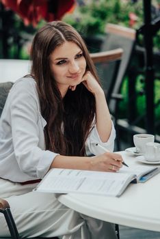 Smiling young dark-haired businesswoman makes notes in a notebook in a cafe on a summer day. Business, e-learning, freelance concept. laptop on a table.