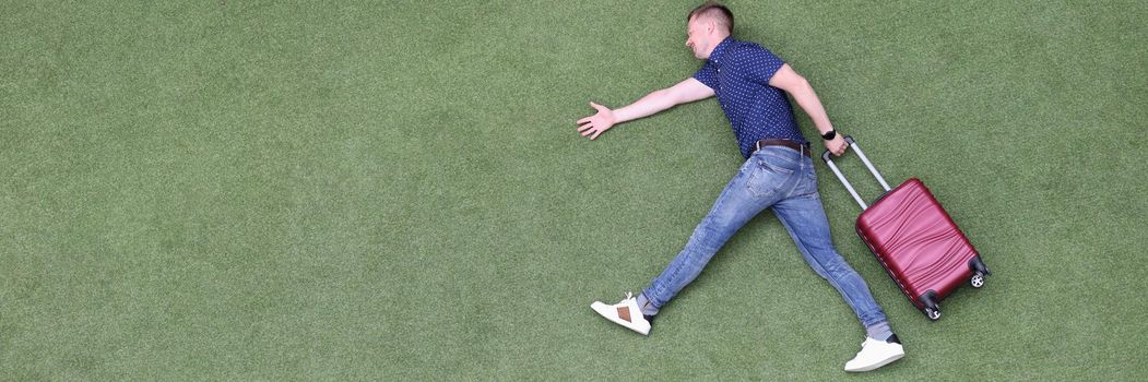 Young man with suitcase walking on green grass top view. Travel dreams concept