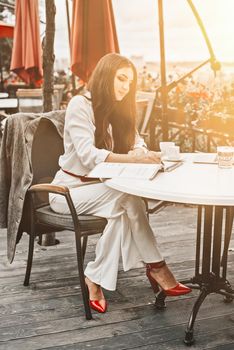 Smiling young dark-haired businesswoman makes notes in a notebook in a cafe on a summer day. Business, e-learning, freelance concept. laptop on a table.