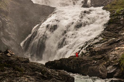 A woman wearing a red dress dancing next to Kjosfossen waterfall in a rocky landscape in Norway