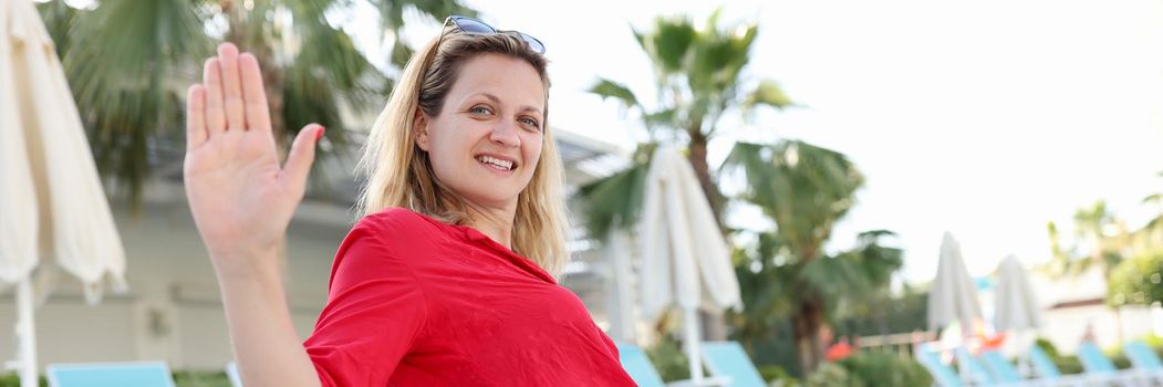 Young woman in red dress sitting on side of pool and waving hand. Rest at resort concept