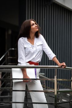 Beautiful young caucasian businesswoman in a white clothes with a laptop and notebook outdoor near business centre.