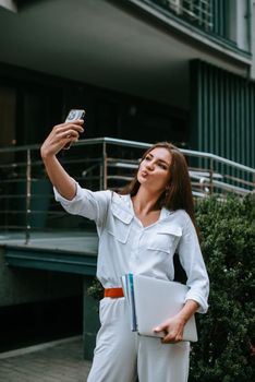 Beautiful young caucasian businesswoman in a white clothes with a laptop and notebook outdoor near business centre.