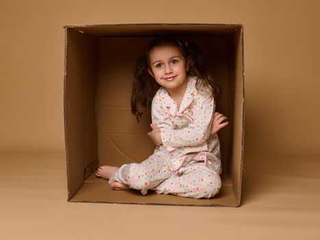 Adorable European little girl in pajamas hugging herself sitting inside a cardboard box and cutely smiling looking at camera, isolated over beige background with copy space for ads