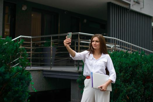 Beautiful young caucasian businesswoman in a white clothes with a laptop and notebook outdoor near business centre.